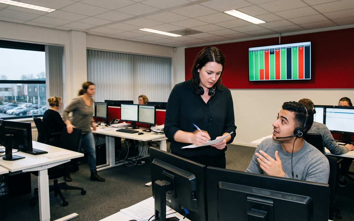 Een klantcontactcentrum, een teamleider met een turflijst tussen de bureaus, een agent met headset kijkt op.