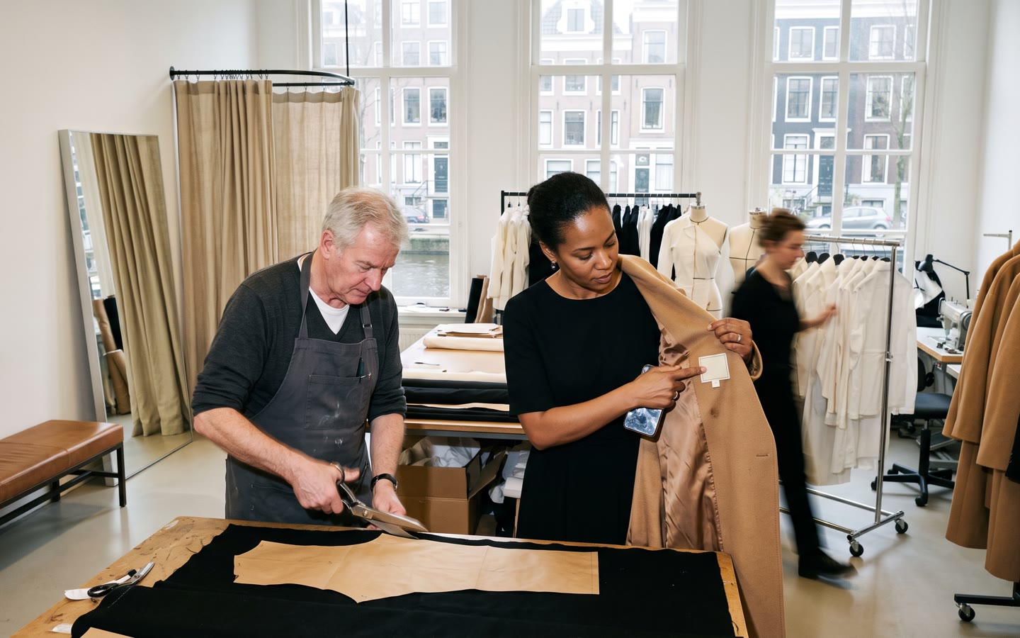 Het atelier van een modehuis aan de gracht, de creatief directeur houdt een jas open bij het label, een patroonmaker aan de snijtafel.