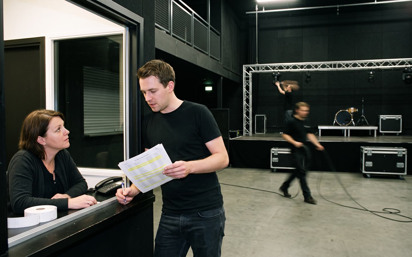 De lege zaal van een poppodium overdag, de programmeur met een ticketlijst bij de kassa, een technicus op het podium.